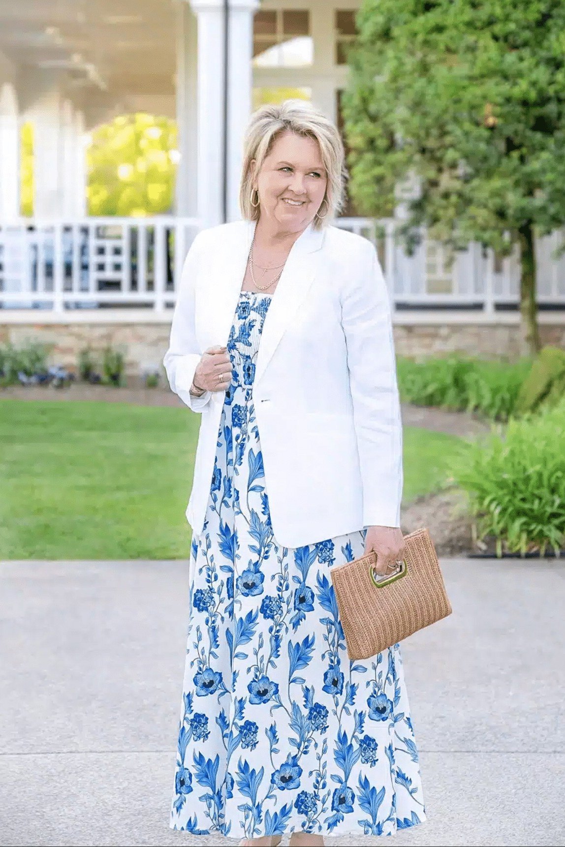 Woman in a blue floral maxi dress layered under a white blazer with a tan clutch - summer dress for women over 50
