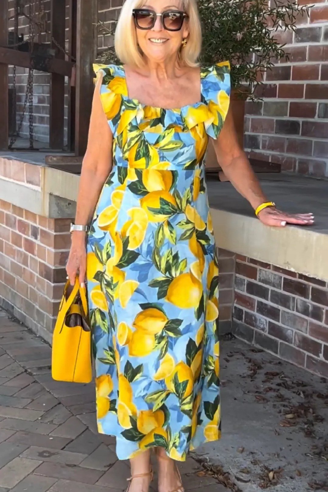 Woman in a yellow lemon-print sleeveless midi dress with a yellow handbag, brick wall - summer dress for women over 50