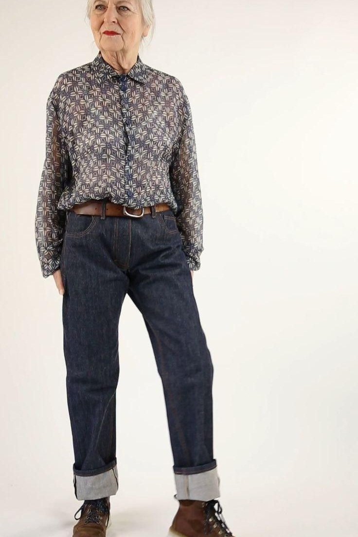 Woman over 60 wearing patterned blouse, dark cuffed jeans, brown boots, studio