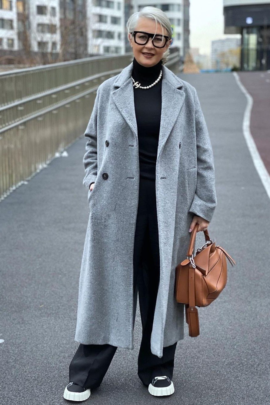 Woman over 60 in a grey long coat over a black outfit with white sneakers