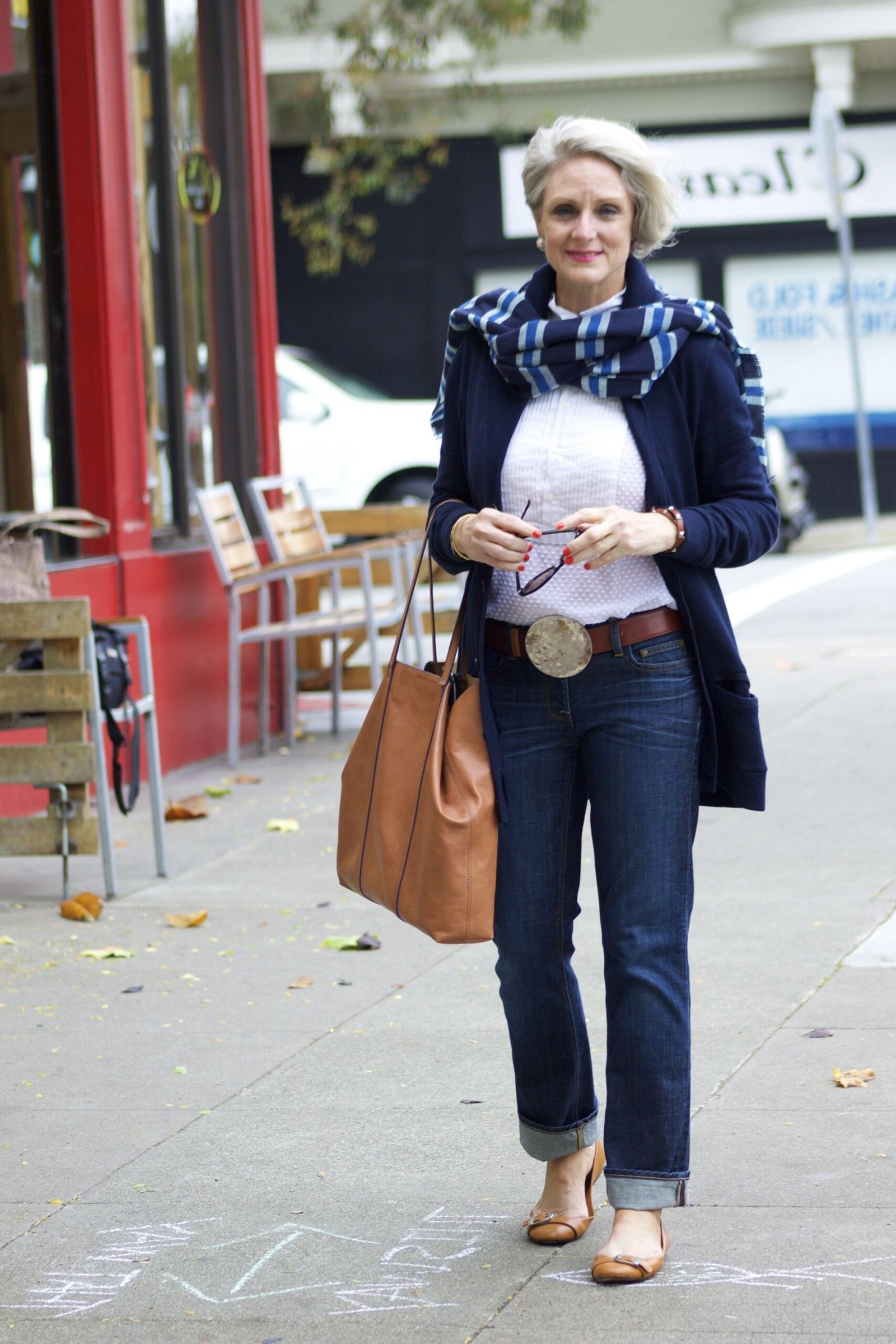 Woman over 60 wearing navy cardigan, white top, patterned scarf, medium jeans, tan bag