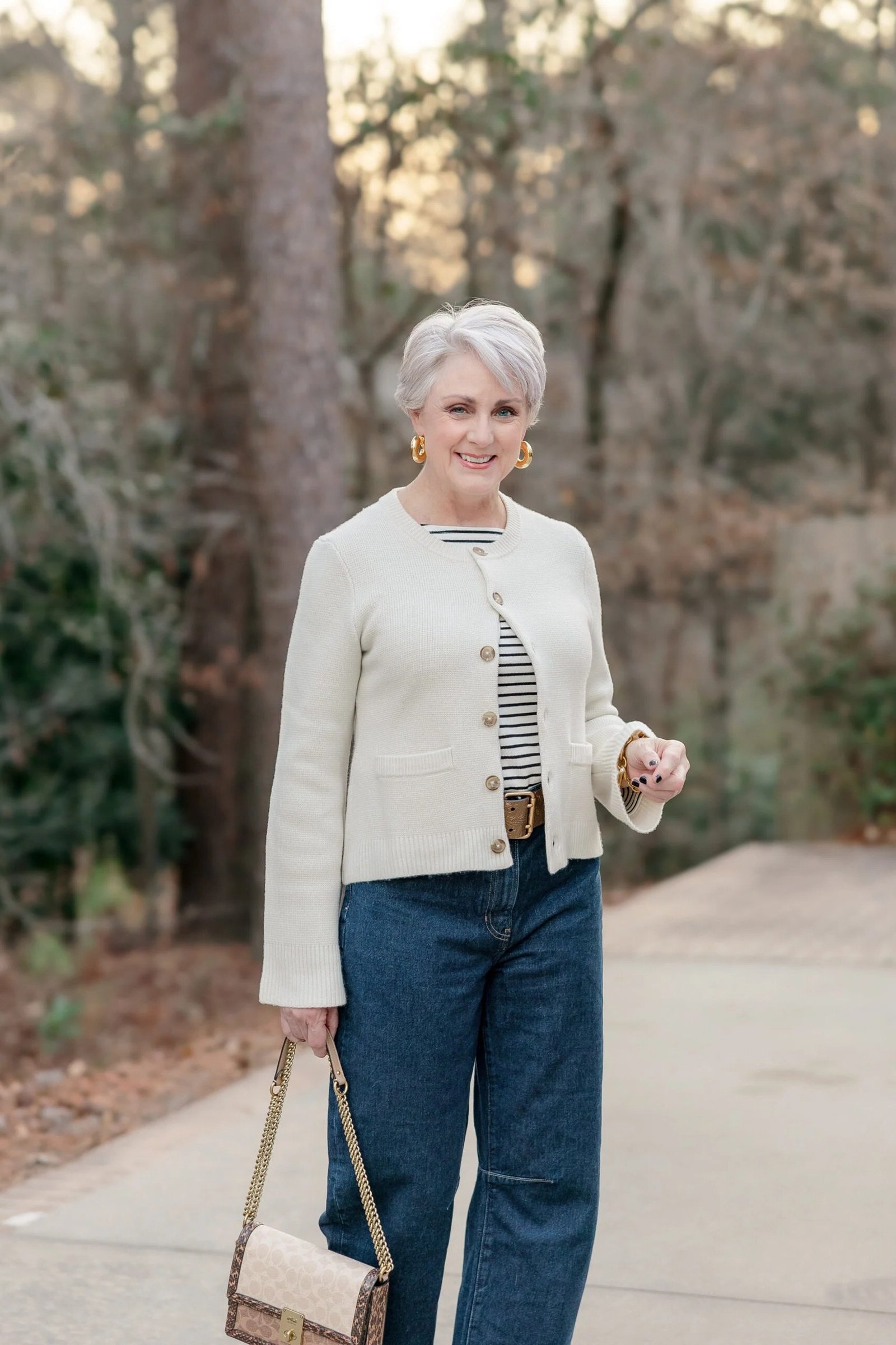 Woman over 60 wearing cream tweed jacket, striped top, wide dark jeans, woven bag