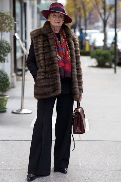 older woman in fur vest with red scarf, black flare pants and burgundy hat, street