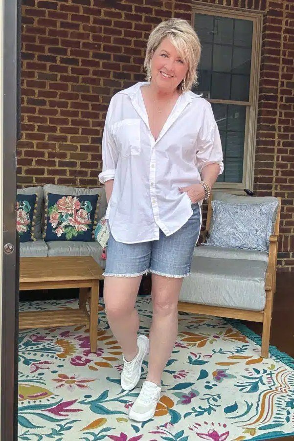 Woman over 50 wearing White button-down shirt with denim shorts and white sneakers on a brick patio.