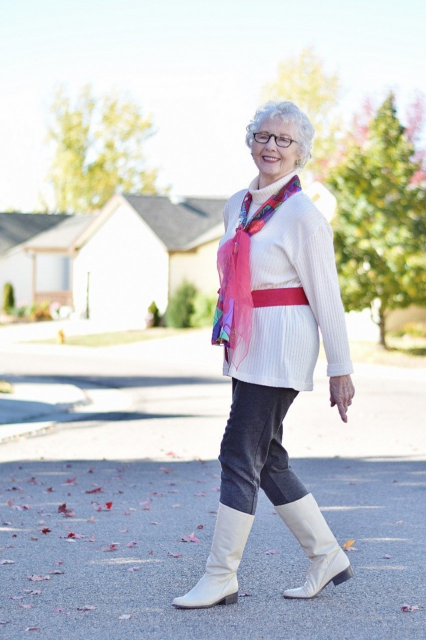 Stylish winter outfit for women over 60: white coat with pink scarf and white boots in a plaza
