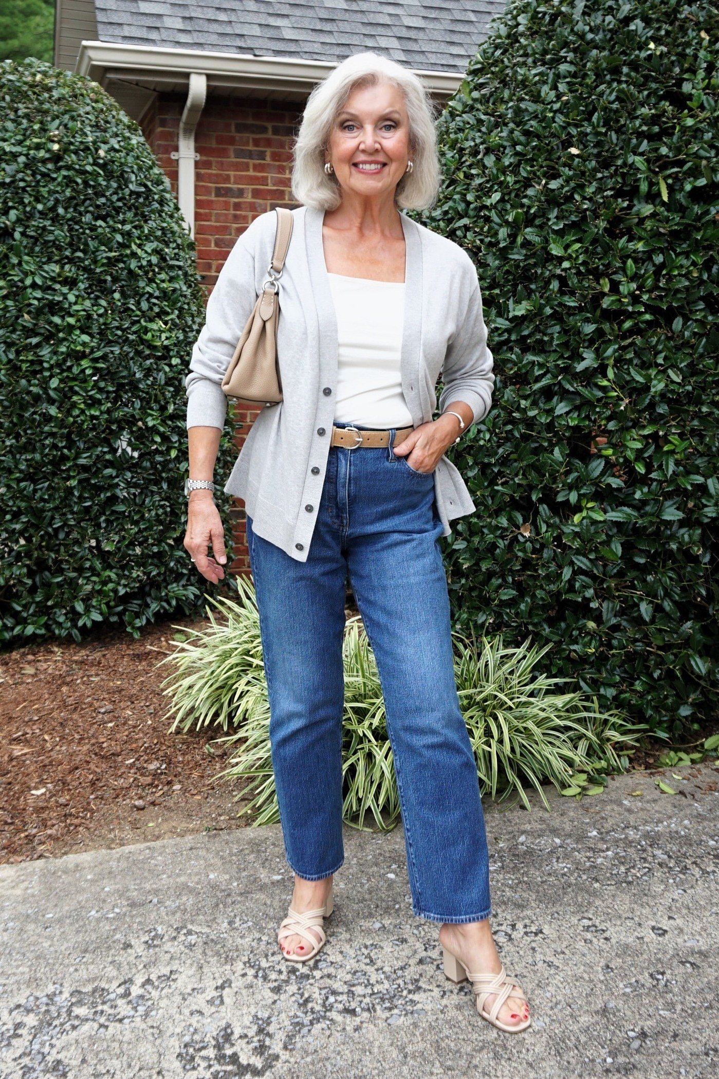Woman over 60 wearing white cardigan, light cropped jeans, sandals, bushes