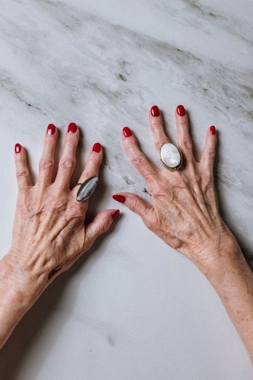 Red glossy nails on elderly hands with rings on marble for women over 70