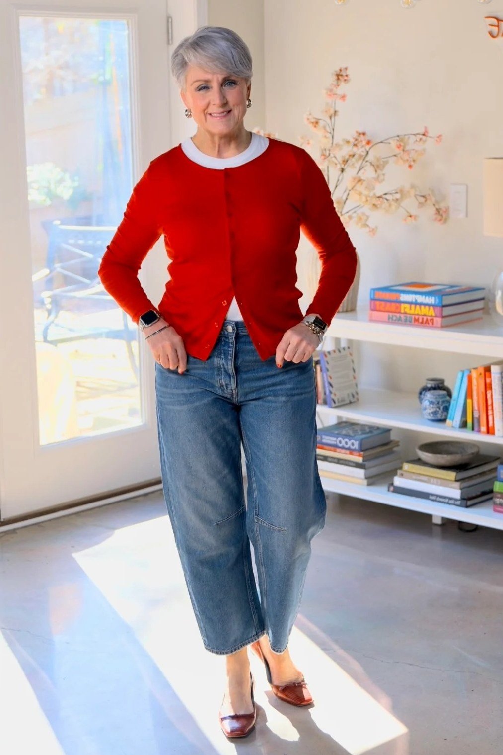 Woman over 60 wearing red cardigan, light wide jeans, red heels, home interior