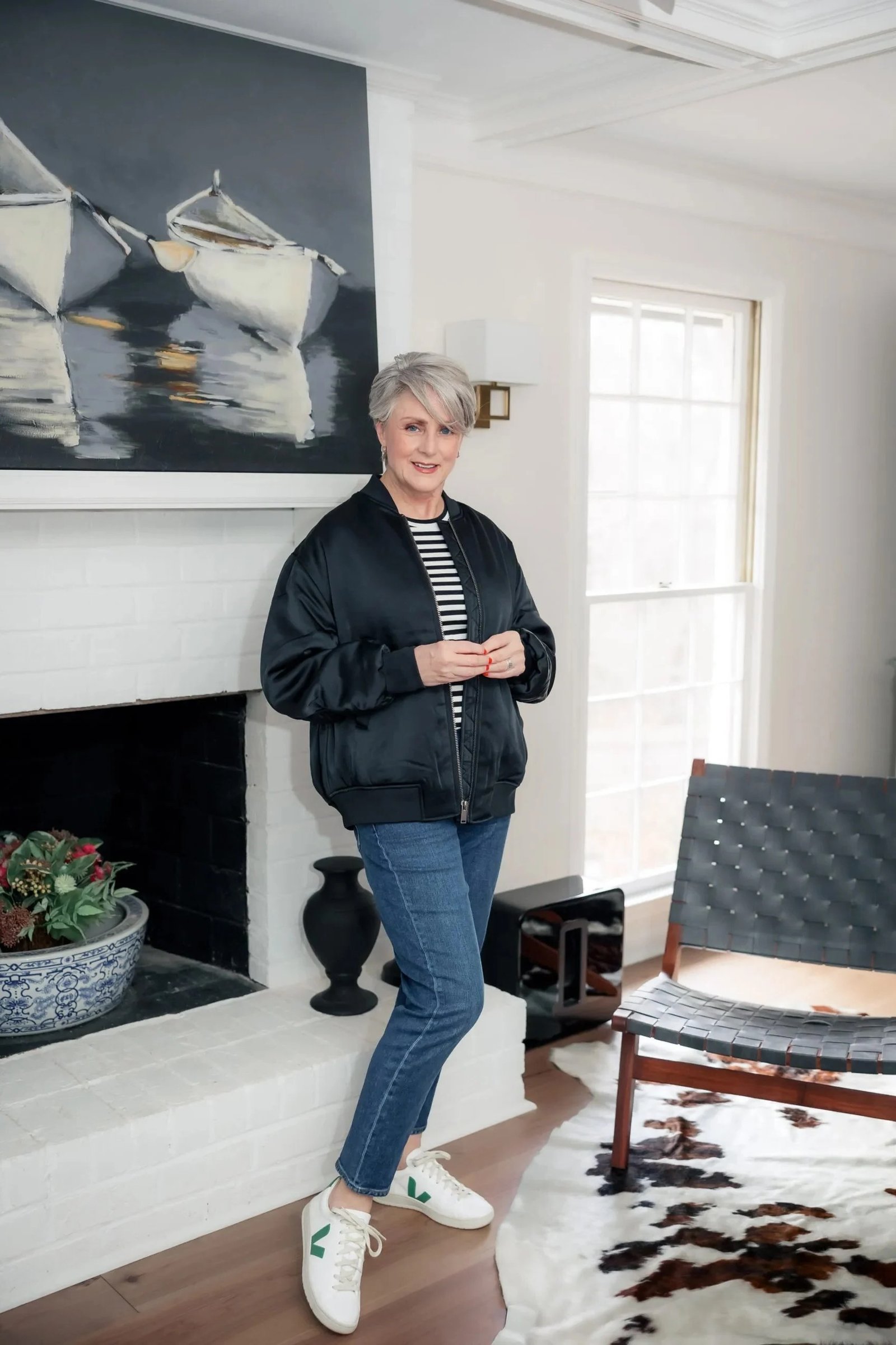 Woman over 50 wearing Black bomber jacket with striped tee, blue jeans and white sneakers by a fireplace.