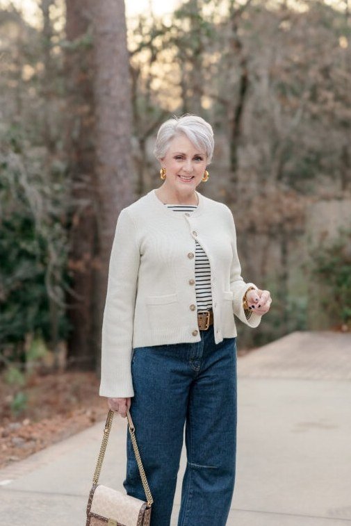 Woman over 60 wearing cream jacket, striped top, wide dark jeans, woven bag, trees