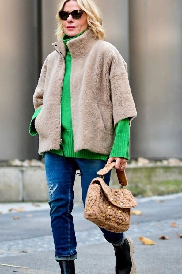 Beige knit poncho over bright green top with blue jeans, cozy October outfit over 50