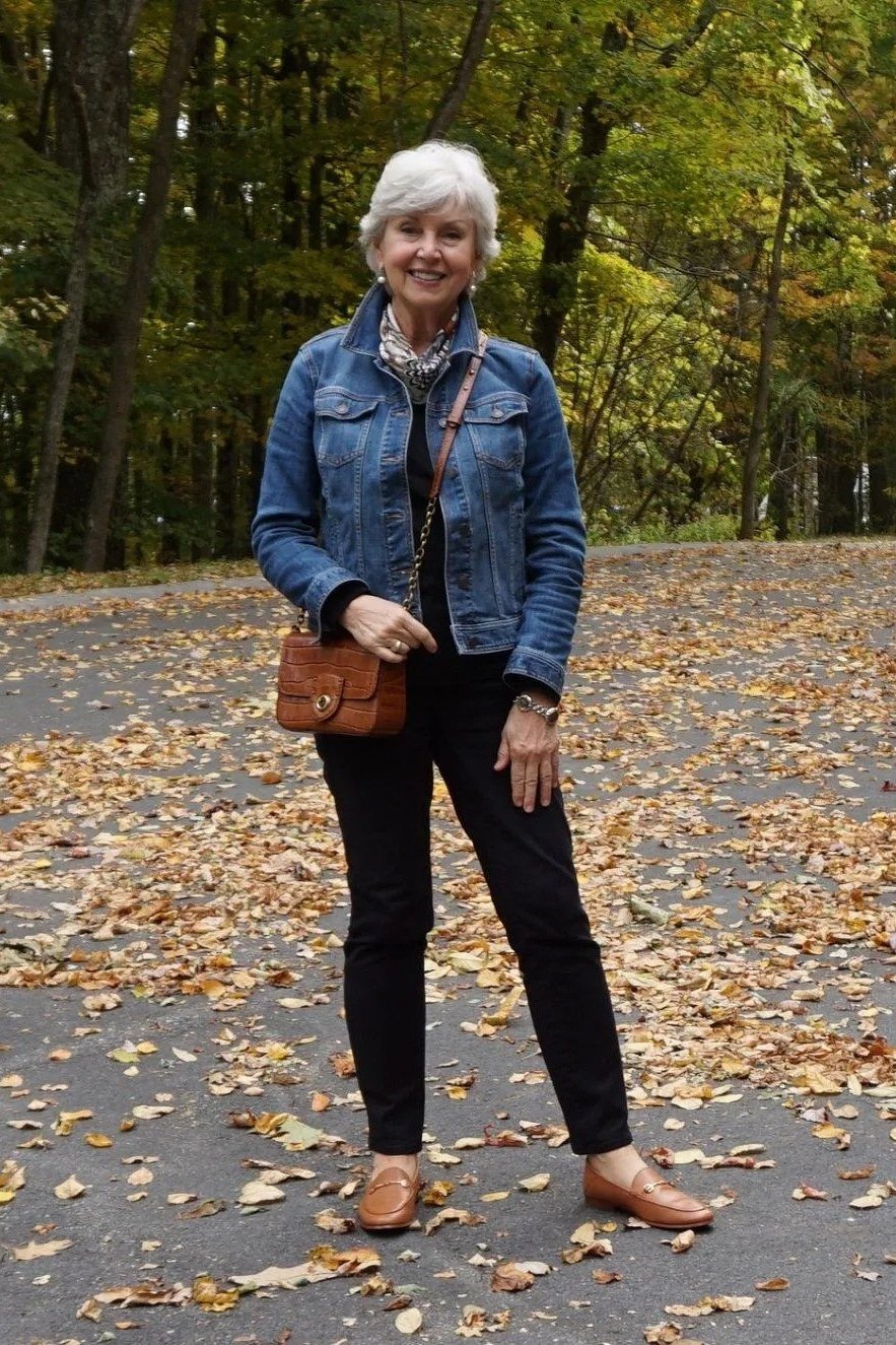 Denim jacket with black trousers and brown crossbody bag on leafy path