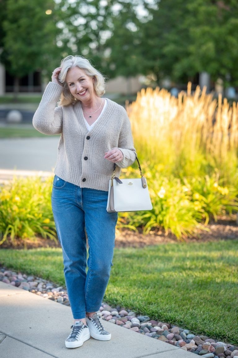 Woman over 60 wearing gray cardigan, white top, medium jeans, white bag, white sneakers, indoors