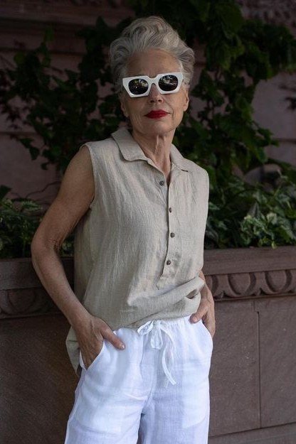 Older woman in a sleeveless beige top and white pants with white sunglasses standing outdoors, an ageless fashion idea for women over 70