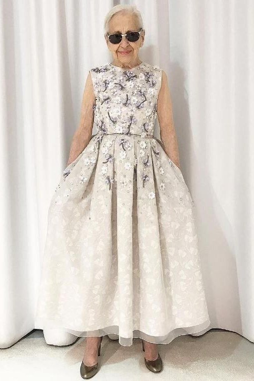 Older woman in a white floral sleeveless gown standing before white curtains, an ageless fashion idea for women over 70