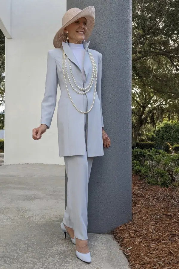 Older woman in a pale-blue tailored suit and wide-brim hat standing outdoors by a stone wall, an ageless fashion idea for women over 70