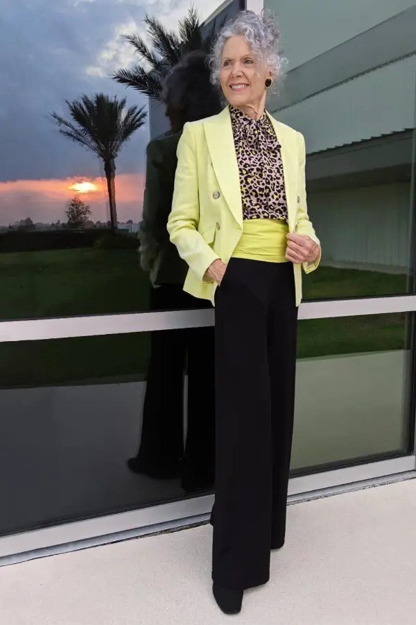 Older woman in a lime-yellow blazer over a leopard-print top with black wide-leg pants at sunset, an ageless fashion idea for women over 70