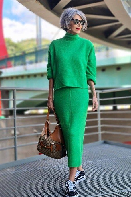 Older woman in a bright-green knit sweater-and-pants set with a brown handbag on a modern walkway, an ageless fashion idea for women over 70