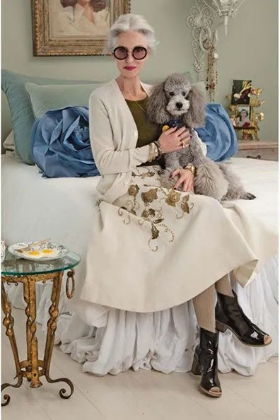 Older woman in a light-blue button-front linen shirtdress with a straw bag in a garden courtyard, an ageless fashion idea for women over 70