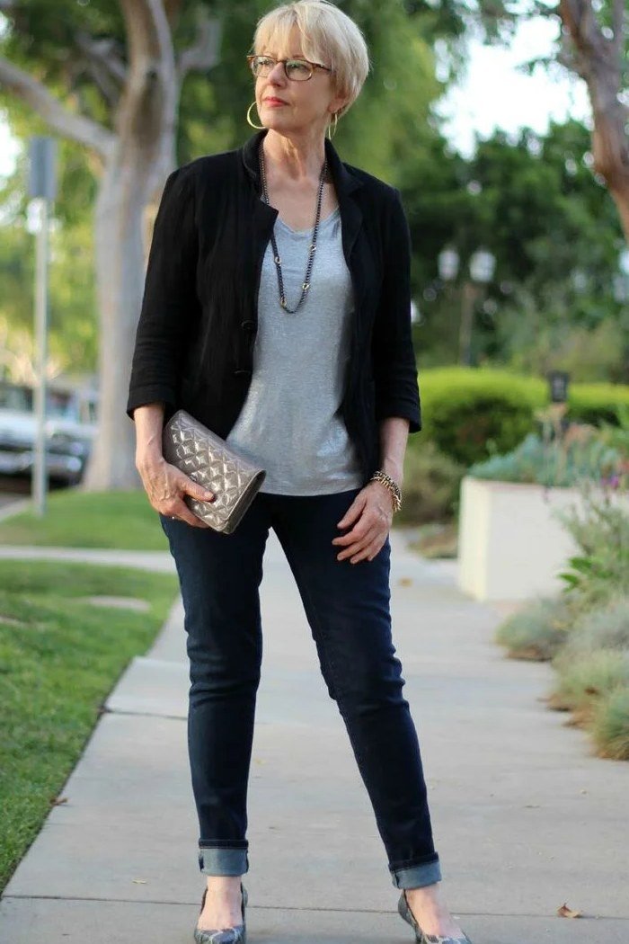 Older woman on a sidewalk in a black blazer over a gray top with dark skinny jeans, heels and a patterned clutch, an ageless fashion idea for women over 70