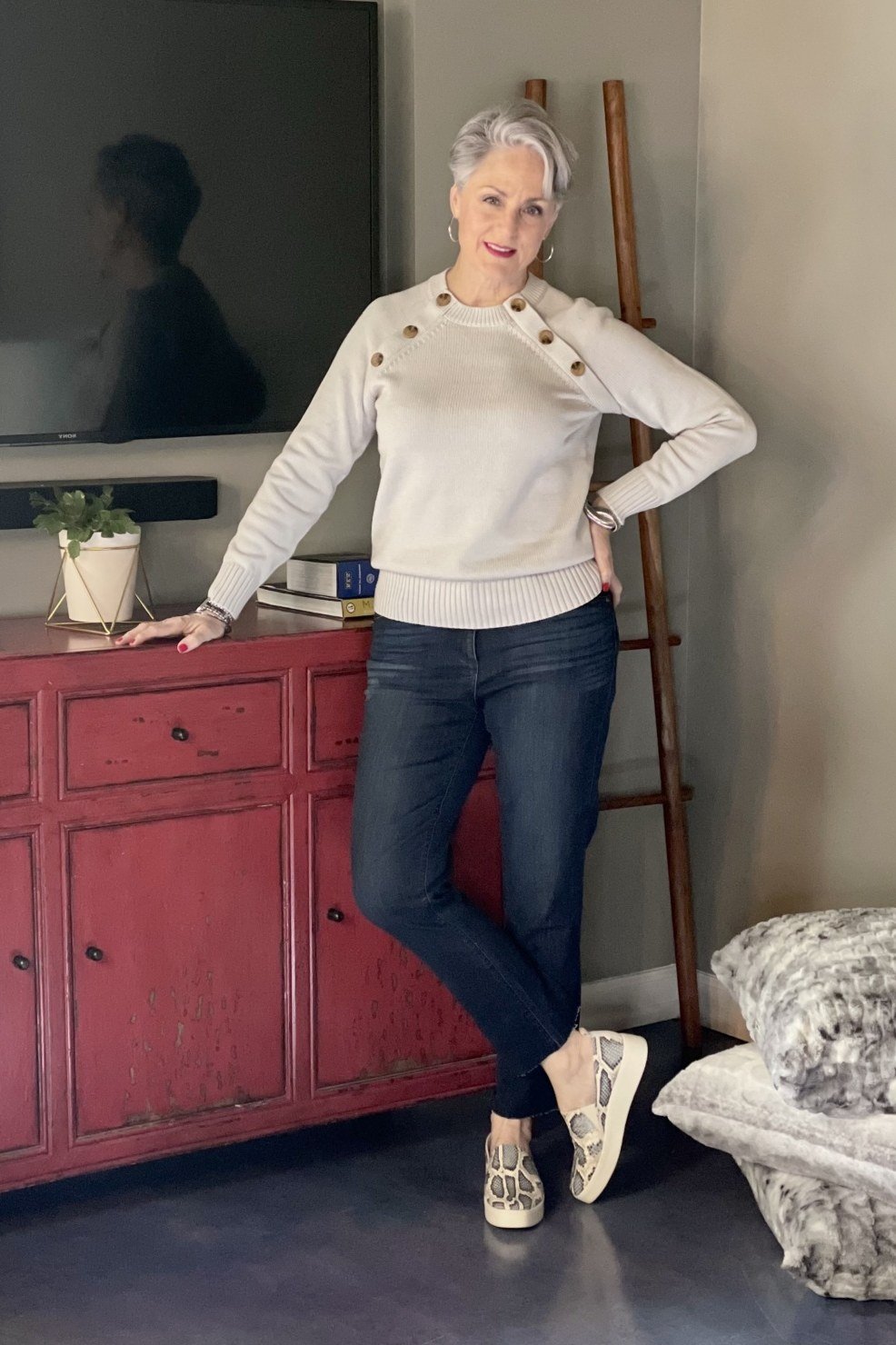 Older woman indoors leaning on a red dresser in a cream knit sweater, dark jeans and beige loafers, an ageless fashion idea for women over 70