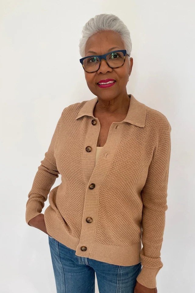 Older Black woman with glasses wearing a tan button-front cardigan over blue jeans, standing in a studio