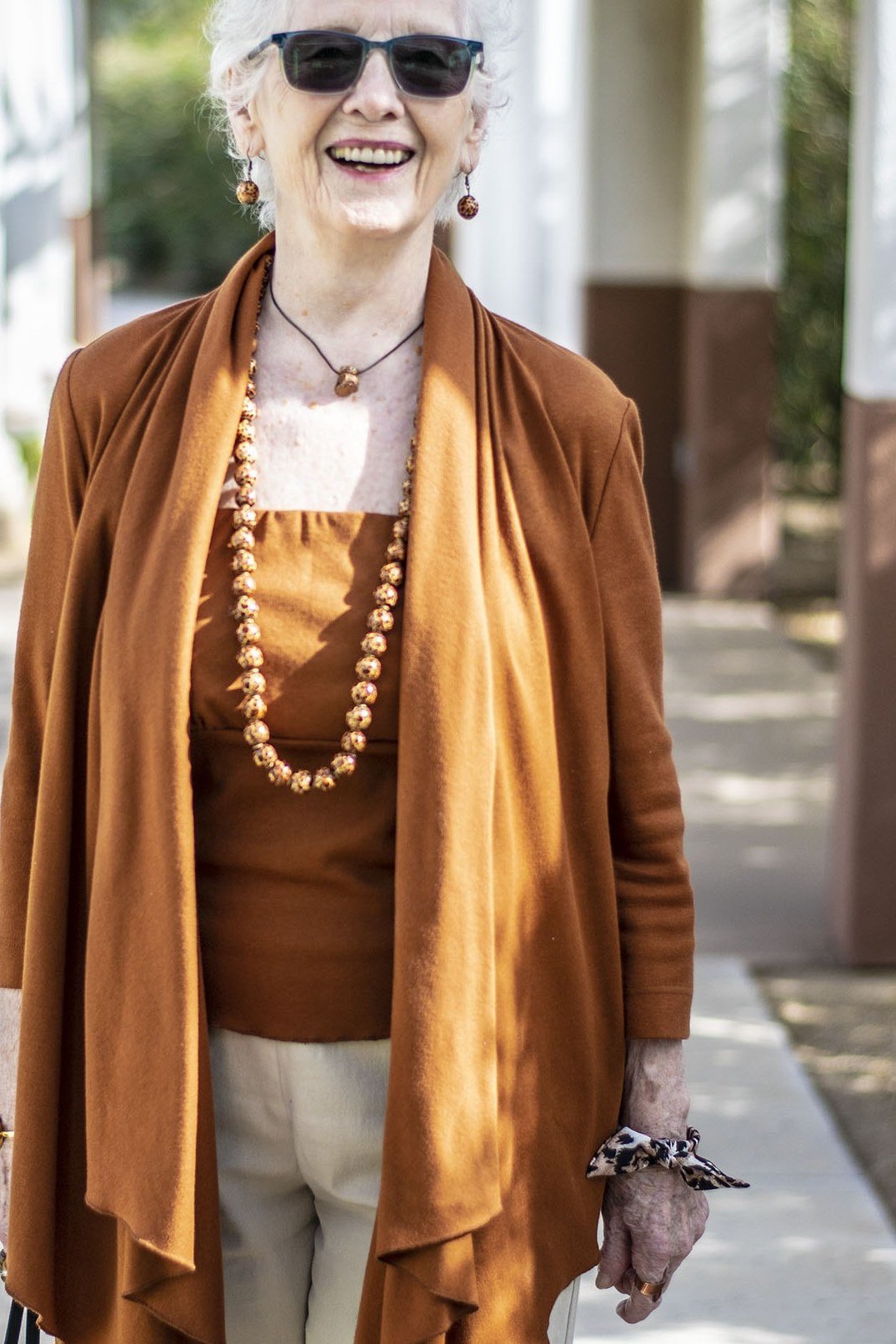 Older woman with sunglasses wearing a rust waterfall cardigan over cream pants with a long beaded necklace