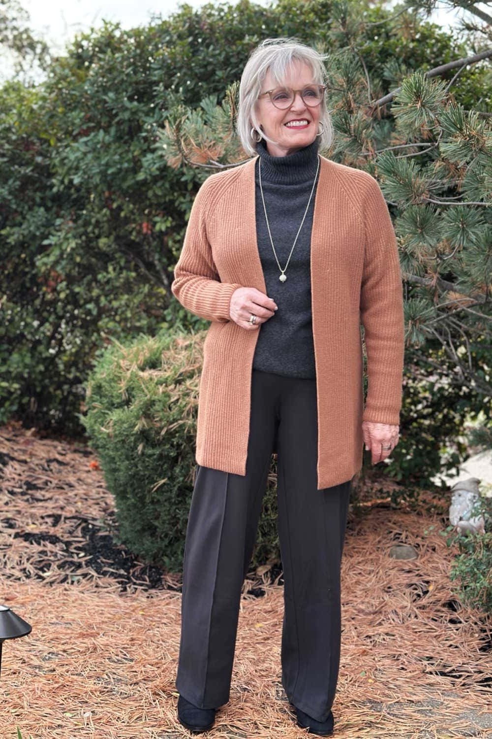 Older woman with glasses wearing a camel long cardigan over a black turtleneck and wide-leg pants outdoors