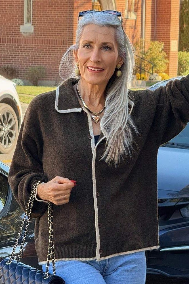 Older woman with long gray hair outdoors wearing a dark brown fleece cardigan with white trim over jeans