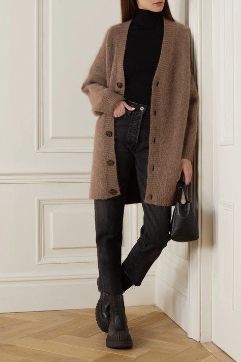 Woman leaning in a doorway wearing a camel long cardigan over a black top, jeans, and boots
