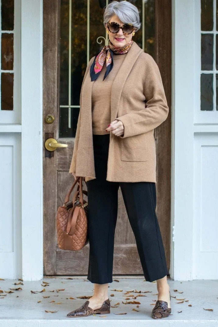 Older woman at a doorway wearing a camel cardigan with a scarf, dark pants, and loafers, carrying a tan bag