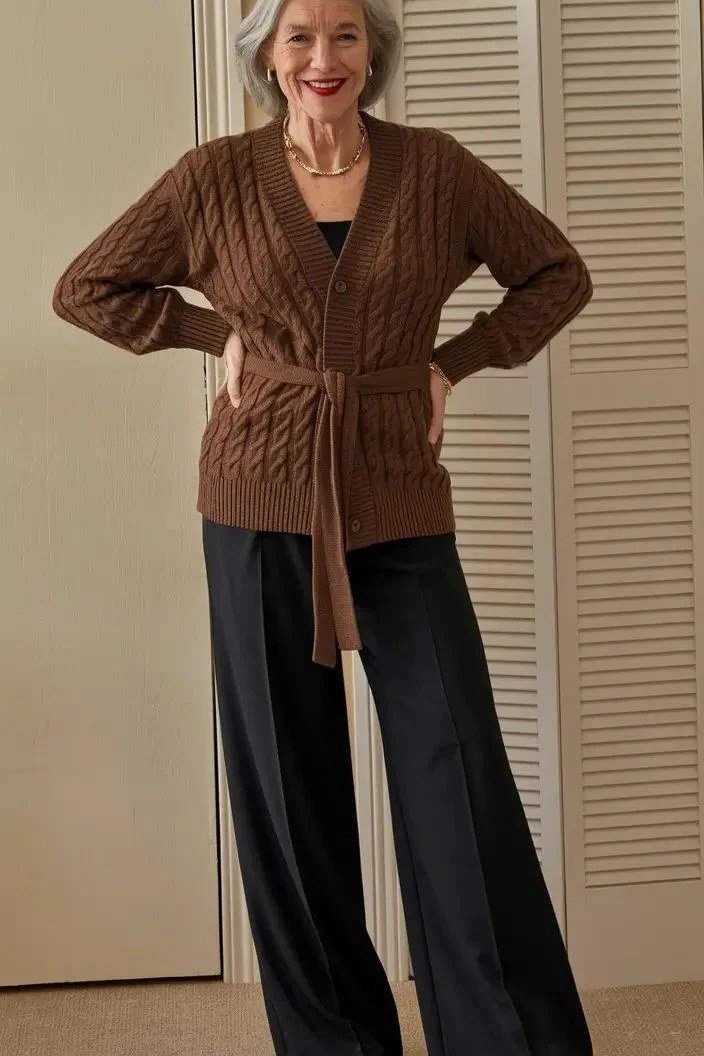 Older woman with hands on hips wearing a dark brown tie-front cable cardigan with black wide-leg pants