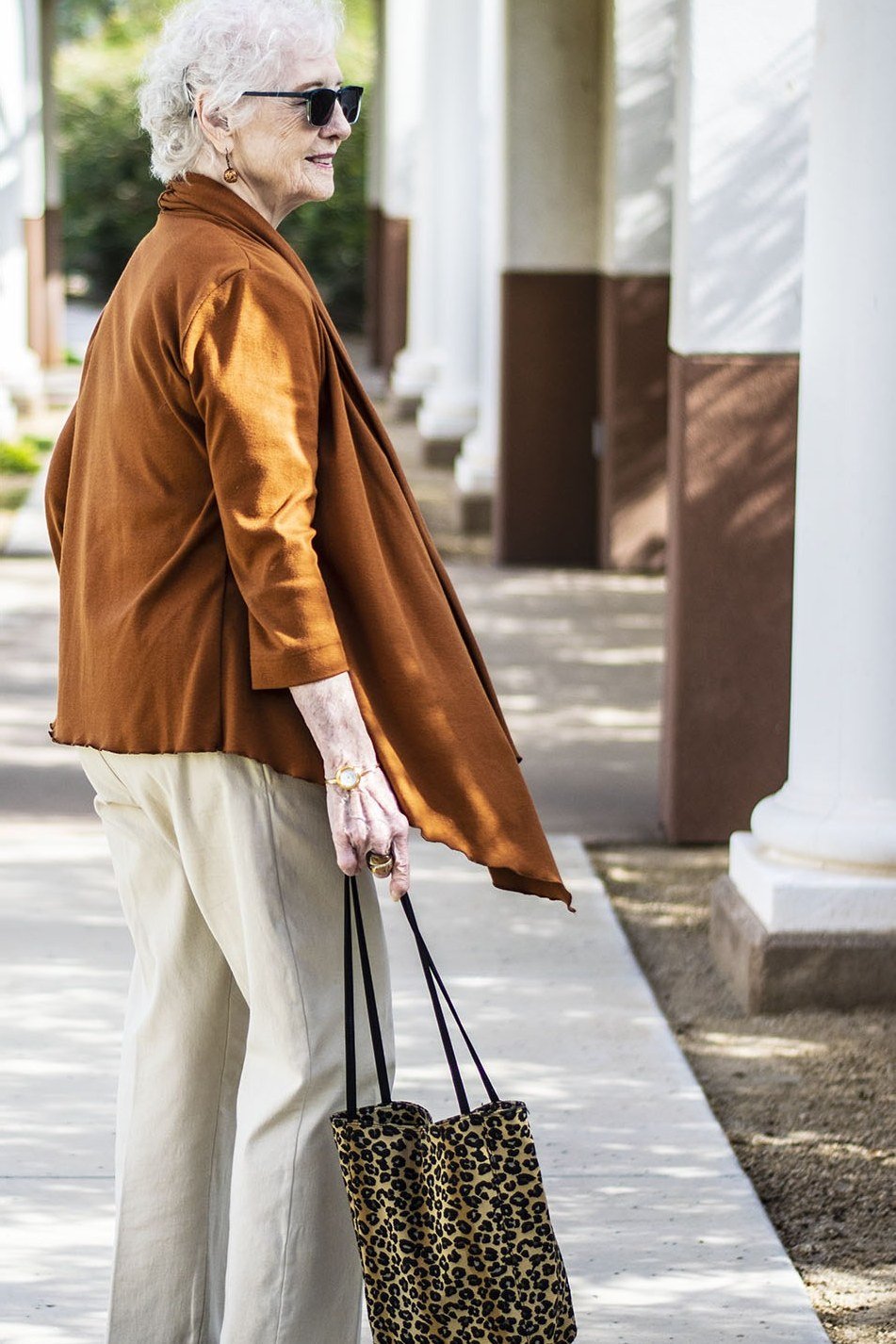 Older woman with sunglasses walking outdoors in a rust-orange cardigan with white pants, carrying a leopard tote
