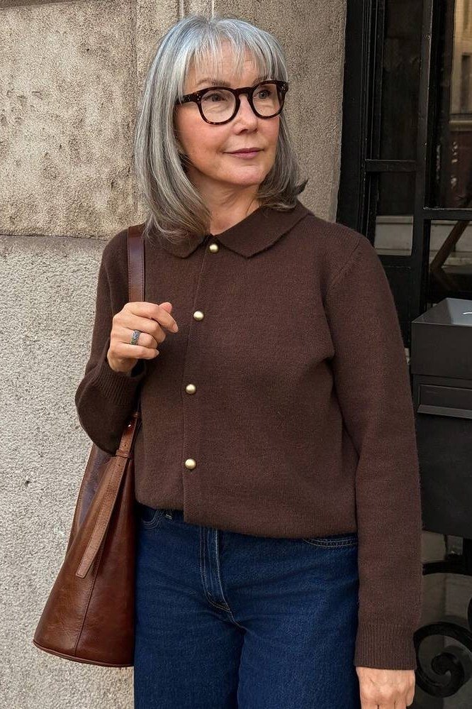 Older woman with glasses wearing a dark brown polo-collar cardigan with jeans and a brown bag