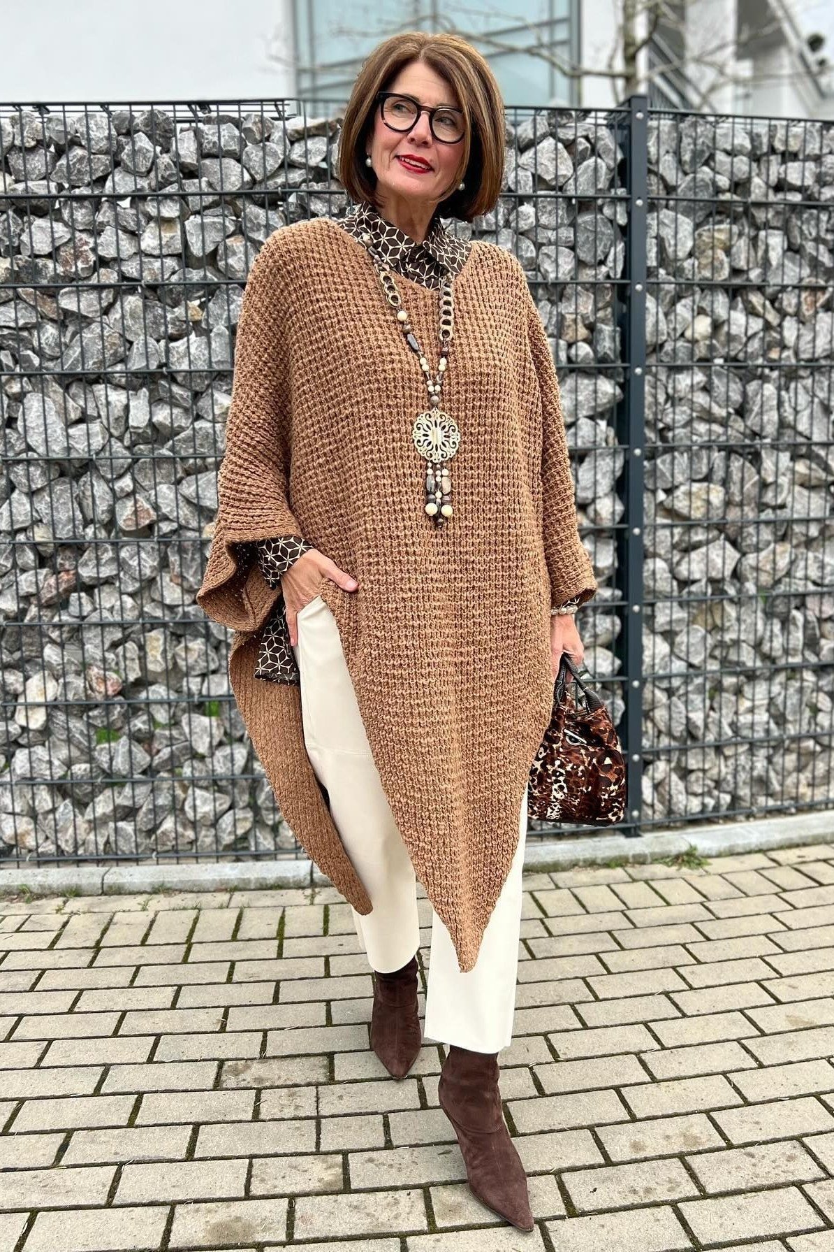 Woman with sunglasses at a stone wall wearing a tan poncho cardigan with white pants