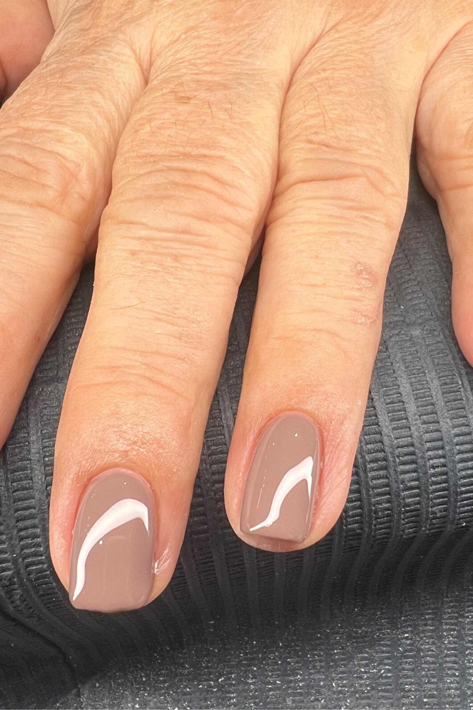 Taupe polish with a thin curved white accent line on each nail. Aged hands on a dark ribbed surface for women over 60