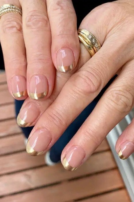 Gold french tips over nude on short nails of mature hands wearing gold rings, on a wooden deck for women over 60