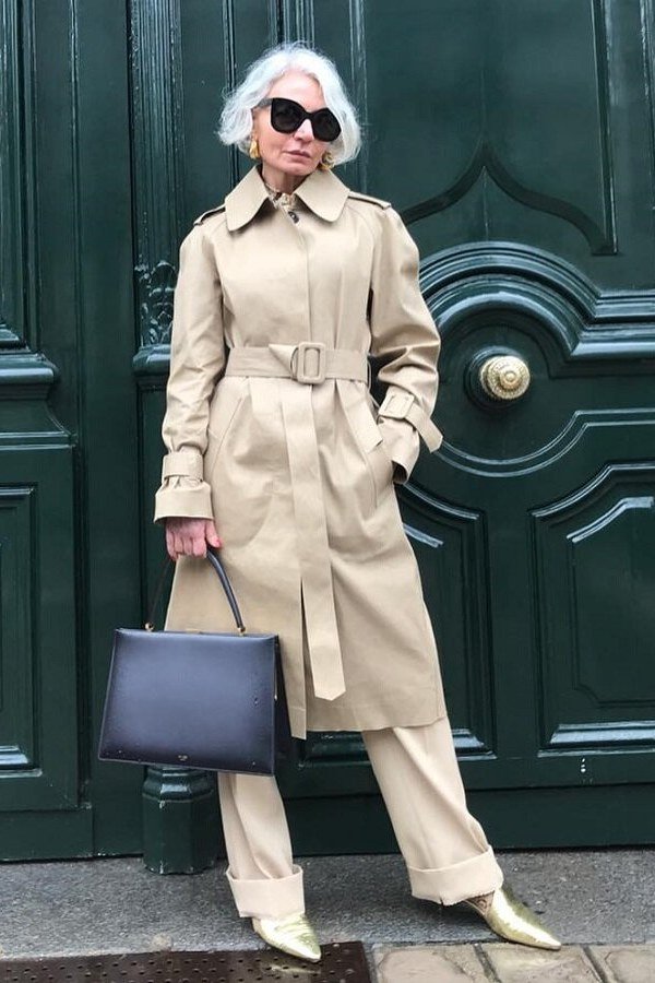 Woman over 60 wearing beige trench coat, white wide trousers, black tote, green door backdrop, sunglasses, classy fall outfit idea