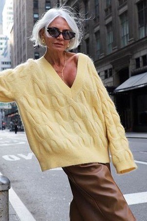 Woman over 60 wearing yellow cable-knit sweater, brown trousers, city street, classy fall outfit idea