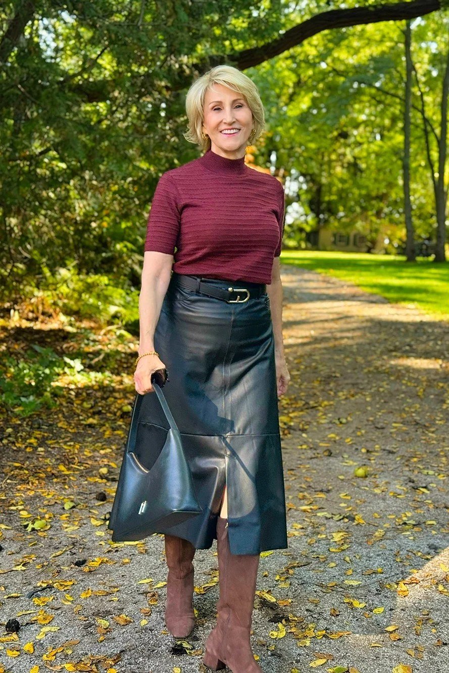 Woman over 60 wearing burgundy turtleneck, black leather midi skirt, brown boots, park, classy fall outfit idea