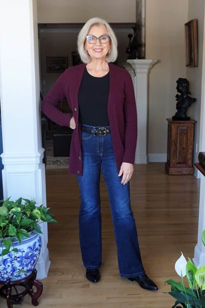 Woman over 60 wearing burgundy cardigan, black top, blue jeans, classy fall outfit idea
