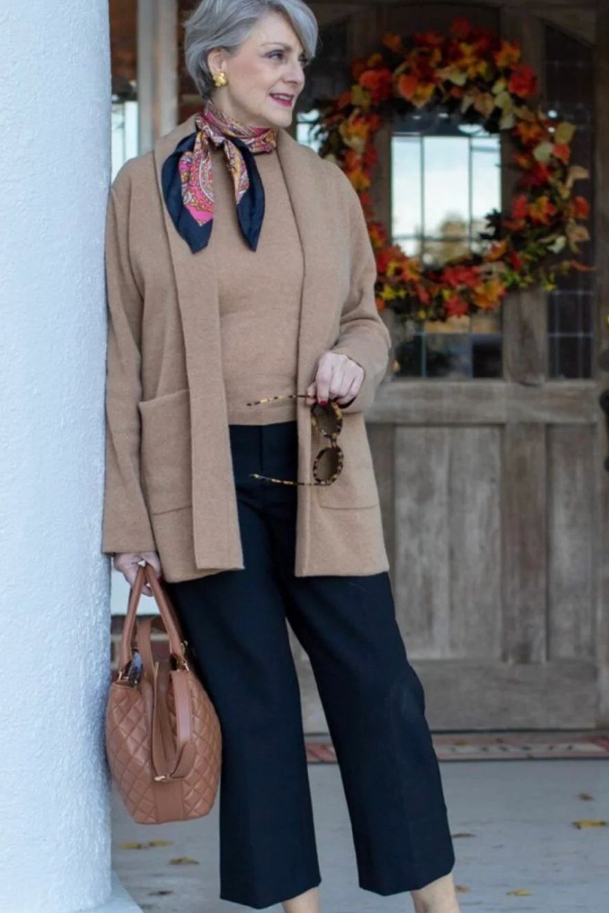 Woman over 60 wearing camel cardigan, patterned scarf, navy cropped trousers, classy fall outfit idea