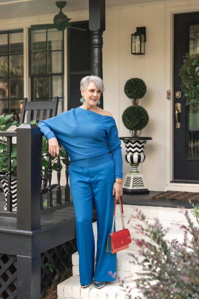 Woman over 50 in cobalt blue one-shoulder top and flared trousers with red handbag