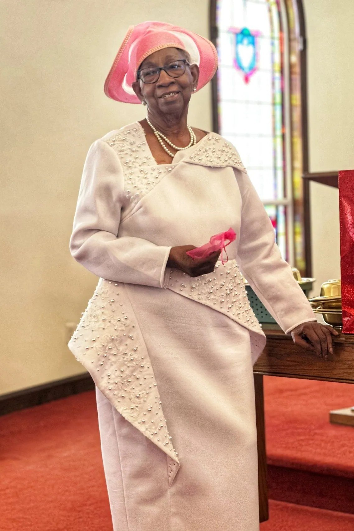 Older woman in cream lace skirt suit with pink hat and gloves in church