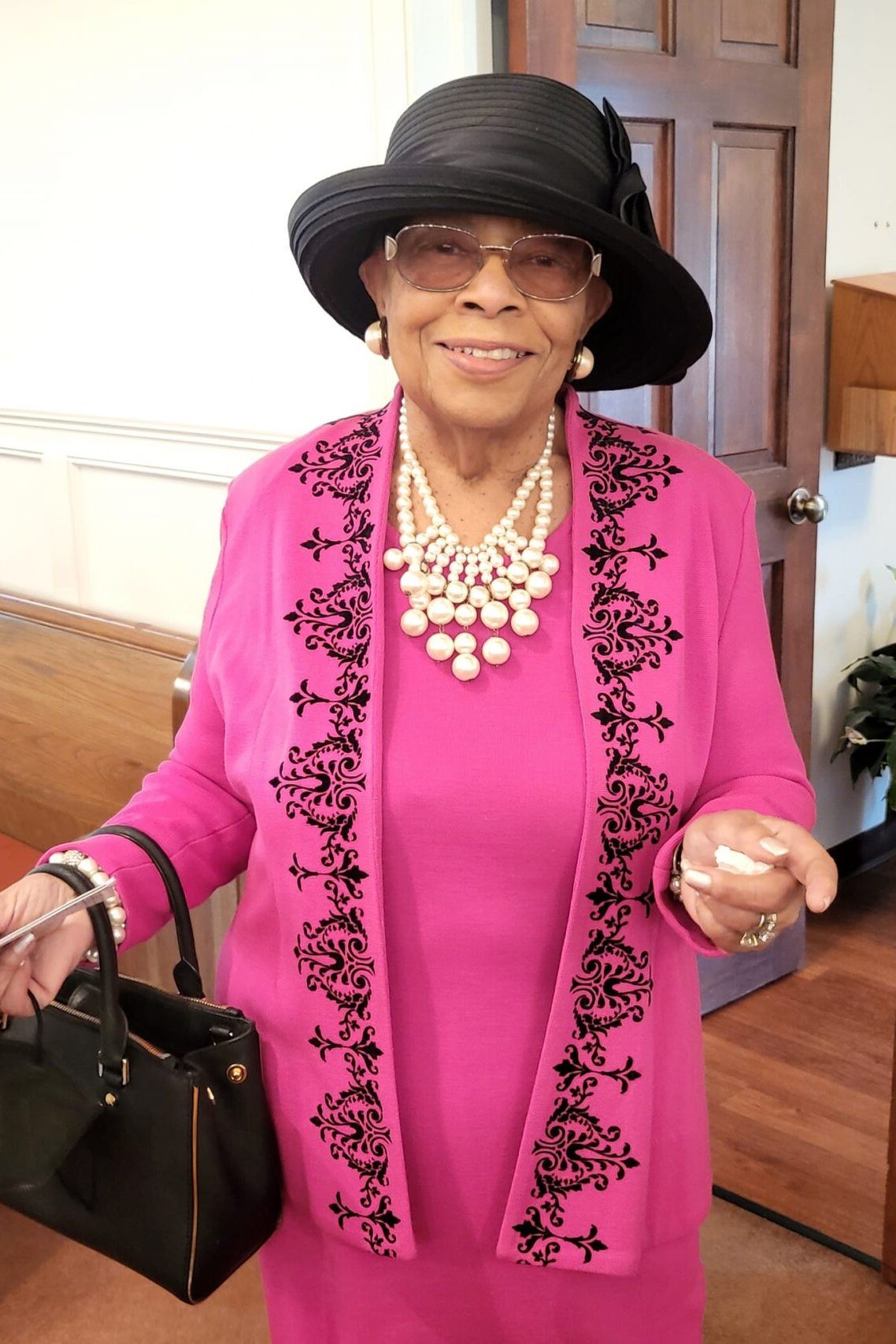 Older woman in fuchsia pink dress with black embroidery and dark hat