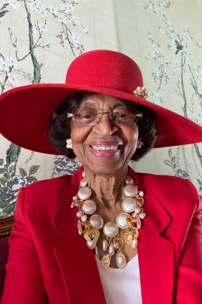 Older woman in red blazer with red wide-brim hat and pearl necklace