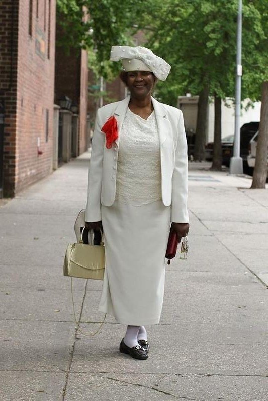 Older woman in white skirt suit with white hat and tan handbag
