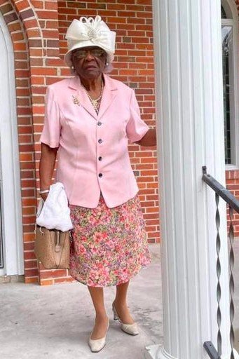 Older woman in pink jacket with floral midi skirt and white hat