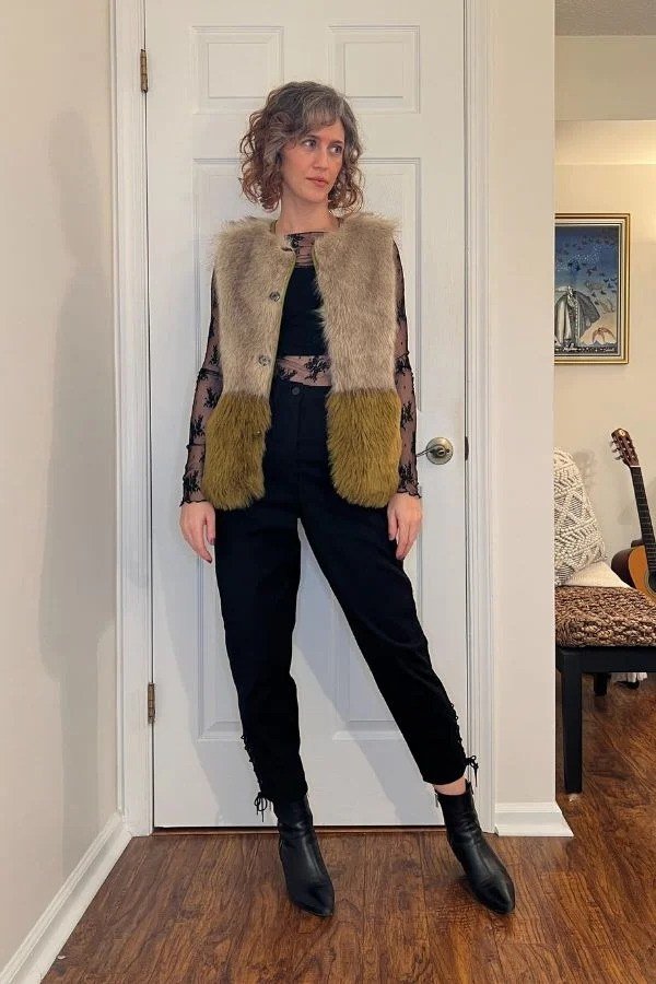 curly-haired woman in her 40s wearing a two-tone faux-fur vest over a black lace top, black ankle-grazing pants and black ankle boots