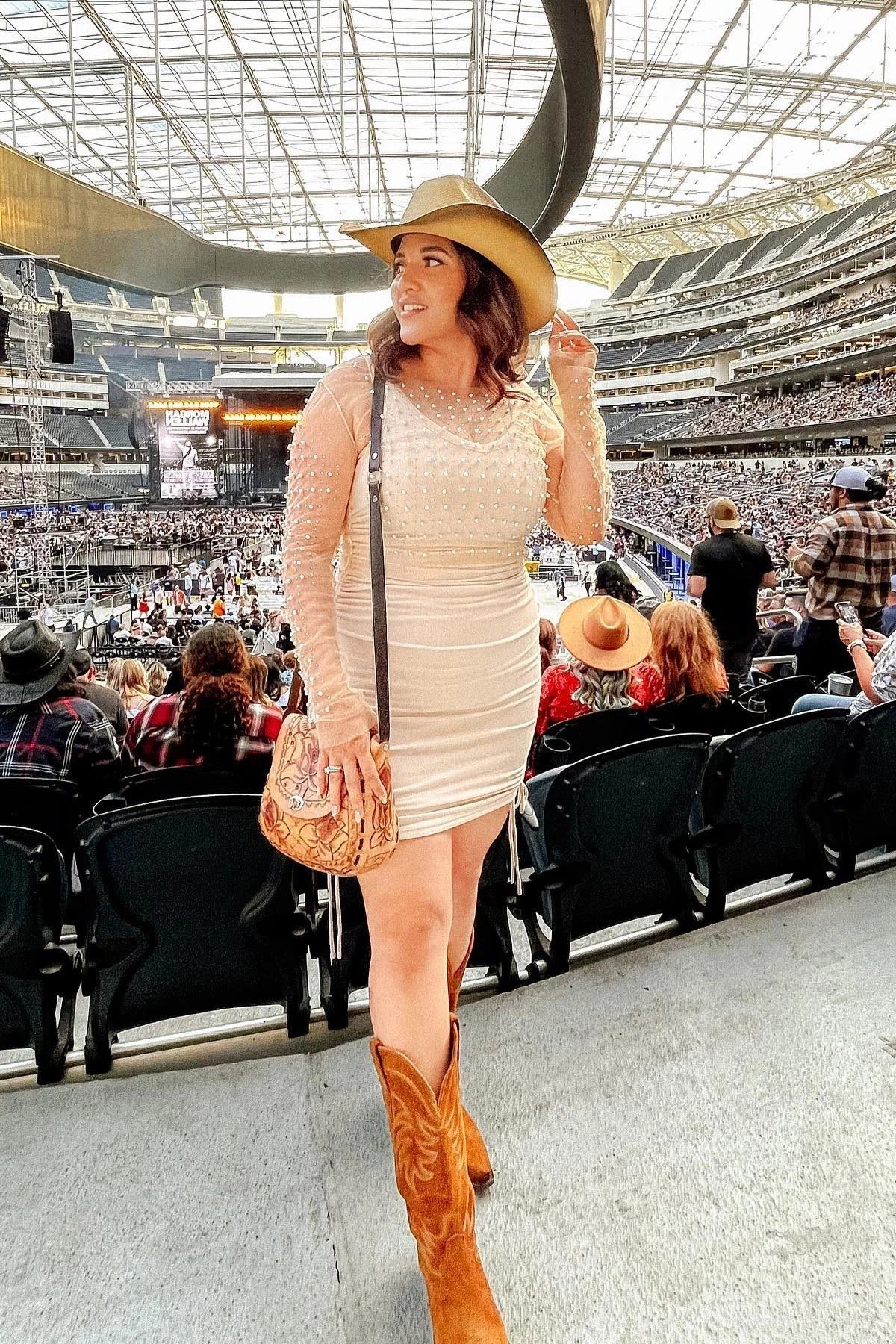 woman in her 40s in stadium seating wearing a fitted ivory sleeveless mini dress, wide-brim straw hat and tall tan cowboy boots